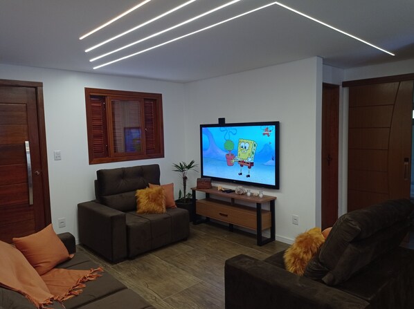Un soggiorno moderno con una televisione a schermo piatto, divani scuri, persiane in legno e un lampadario.