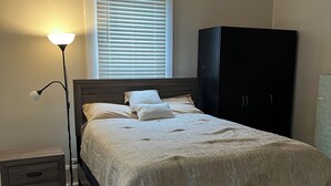 A bedroom with a wooden bed, a nightstand, a lamp, a window with blinds, and a dark wardrobe.