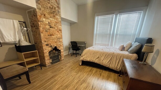 A bedroom with a brick fireplace, wooden flooring, a bed with a headboard, a desk with a chair, and a window with blinds.
