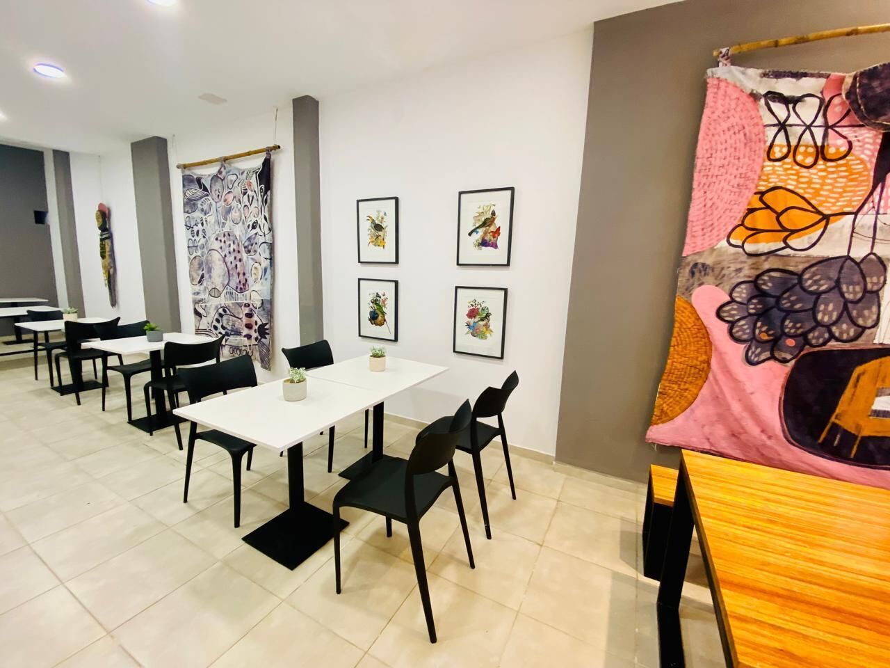 A dining area with white tables and black chairs, framed artwork on the walls, and a colorful tapestry on the right.
