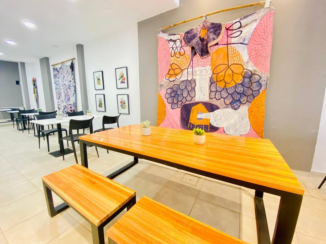A dining area with wooden tables and benches, a colorful tapestry on the wall, and framed pictures on the wall.