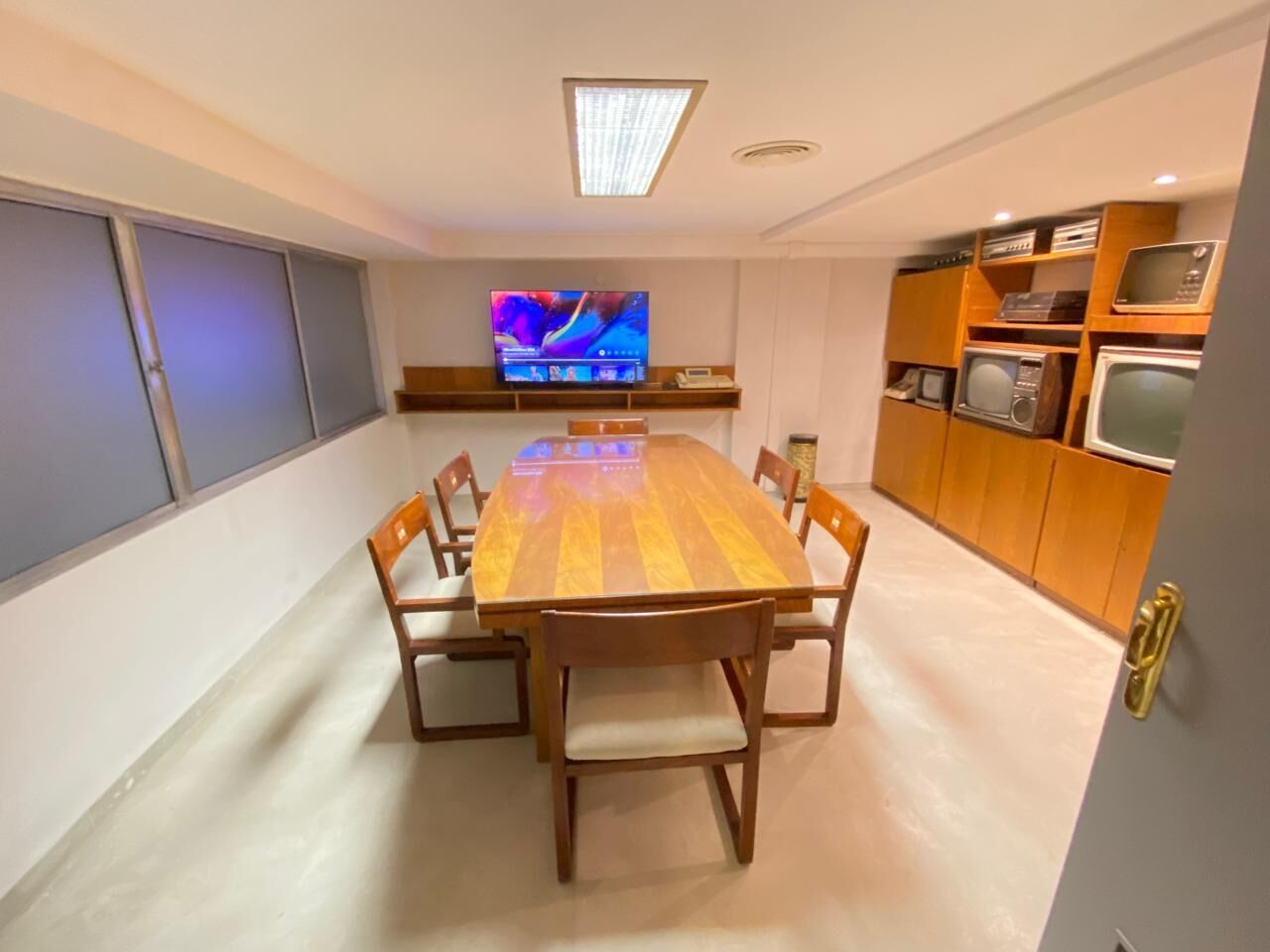A dining area with a wooden table and chairs, a television on the wall, and a kitchen area with wooden cabinets and appliances.