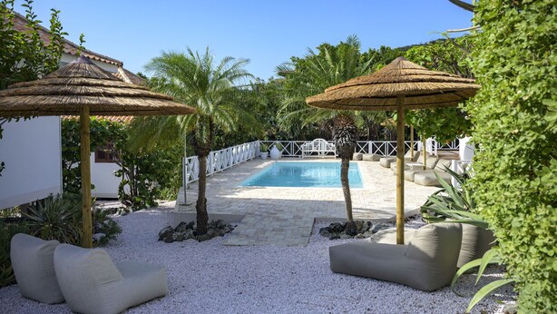 Un área de piscina con una piscina azul cristalina, rodeada de piedrecitas blancas y equipada con dos tumbonas y dos sombrillas de paja.