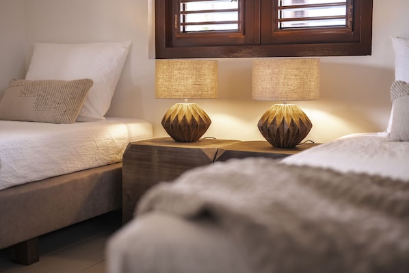 Un dormitorio con dos camas, cada una con un almohadón blanco y una manta texturizada. Entre las camas hay dos mesitas de noche con lámparas.