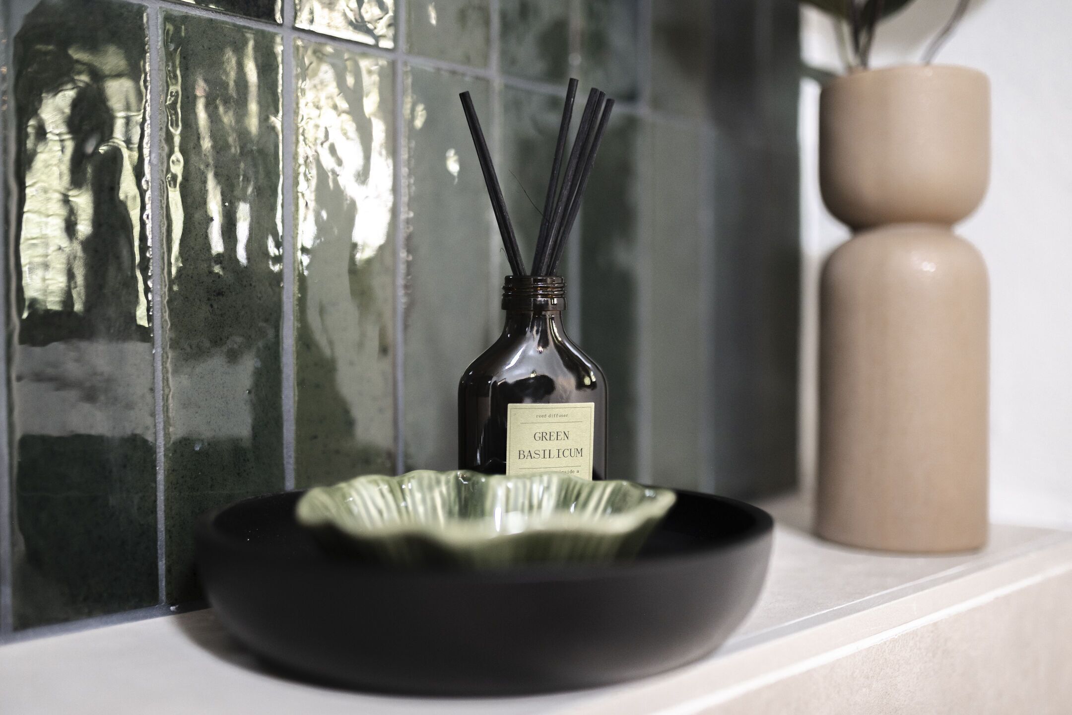 Een zwarte kom op een witte ondergrond met een fles met de tekst 'GREEN BASILICUM' en rietverspreiders voor een betegelde muur.