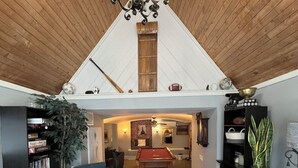 A cozy loft-style living room with a high ceiling, wooden beams, a pool table, and a chandelier. - The Castle - 4-bedroom house with AC, WiFi in Grey County (Feversham)