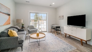 A modern living room with a grey sofa, a round wooden coffee table, a white cabinet with a flat-screen TV, and a patterned rug.