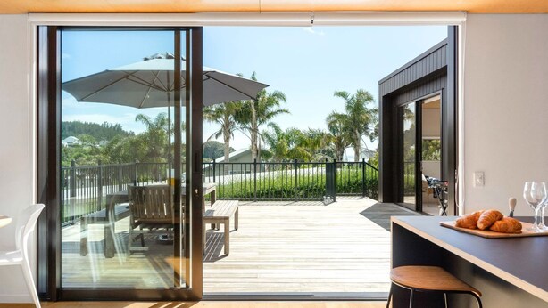 Une cuisine moderne avec un plan de travail en bois, un coin repas avec un banc et un parasol, et une vue sur une terrasse en bois et des palmiers.
