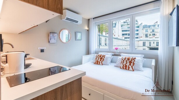 A modern hotel room with a bed, bedside table, and a view of the city through large windows. - De Montescal - Cozy A/C small Studio (Paris)