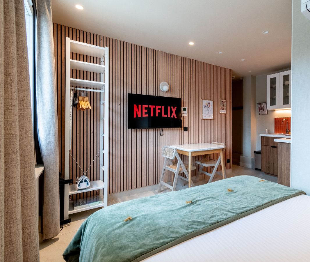 Ein modernes Hotelzimmer mit Bett, Schreibtisch, Stuhl, Wandfernseher und einer Küchenzeile.