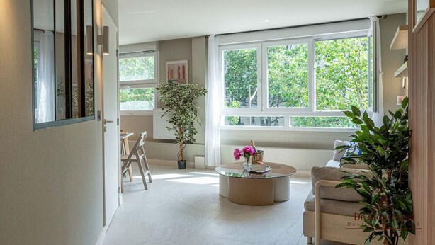 A modern living room with a large window, a round coffee table, a sofa, and a potted plant. - De Montescal - Hot Tub A/C small appartment (Paris)
