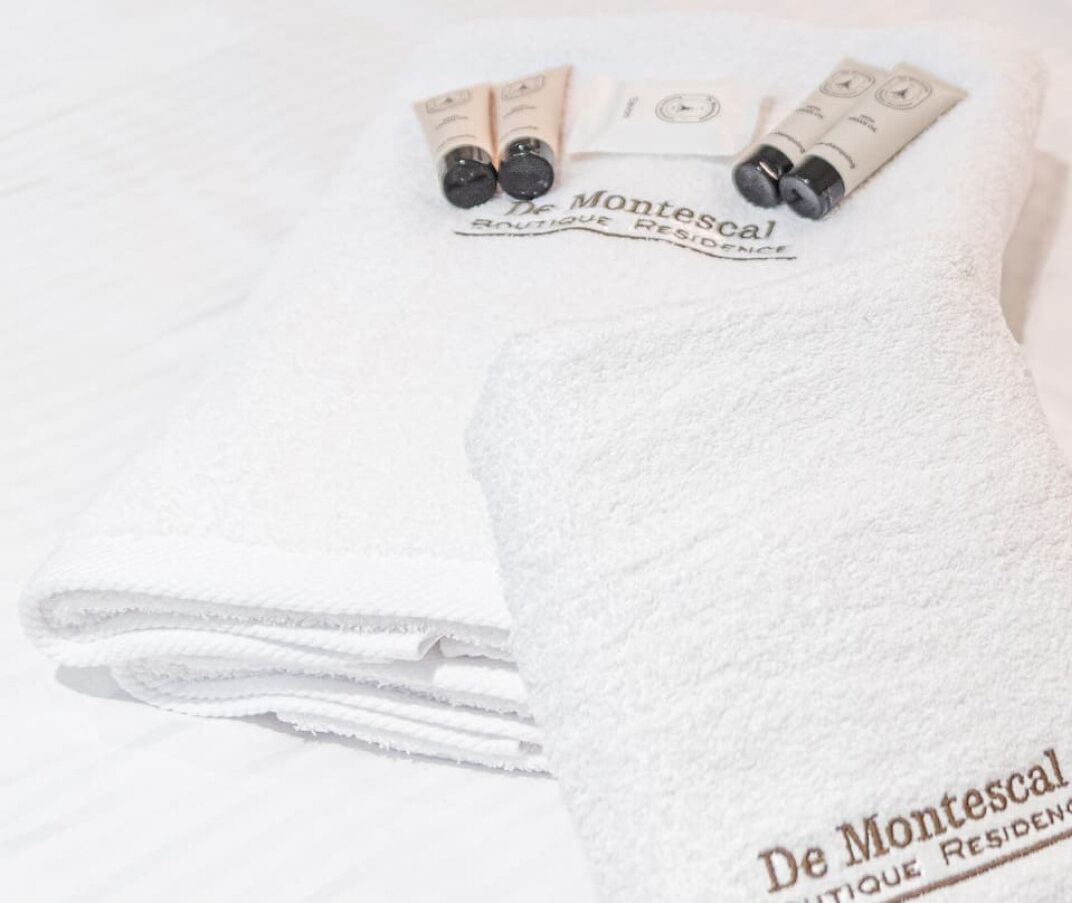Een witte handdoek met de tekst 'De Montescal Boutique Residence' erop, gelegd op een bed met een witte lakenset.