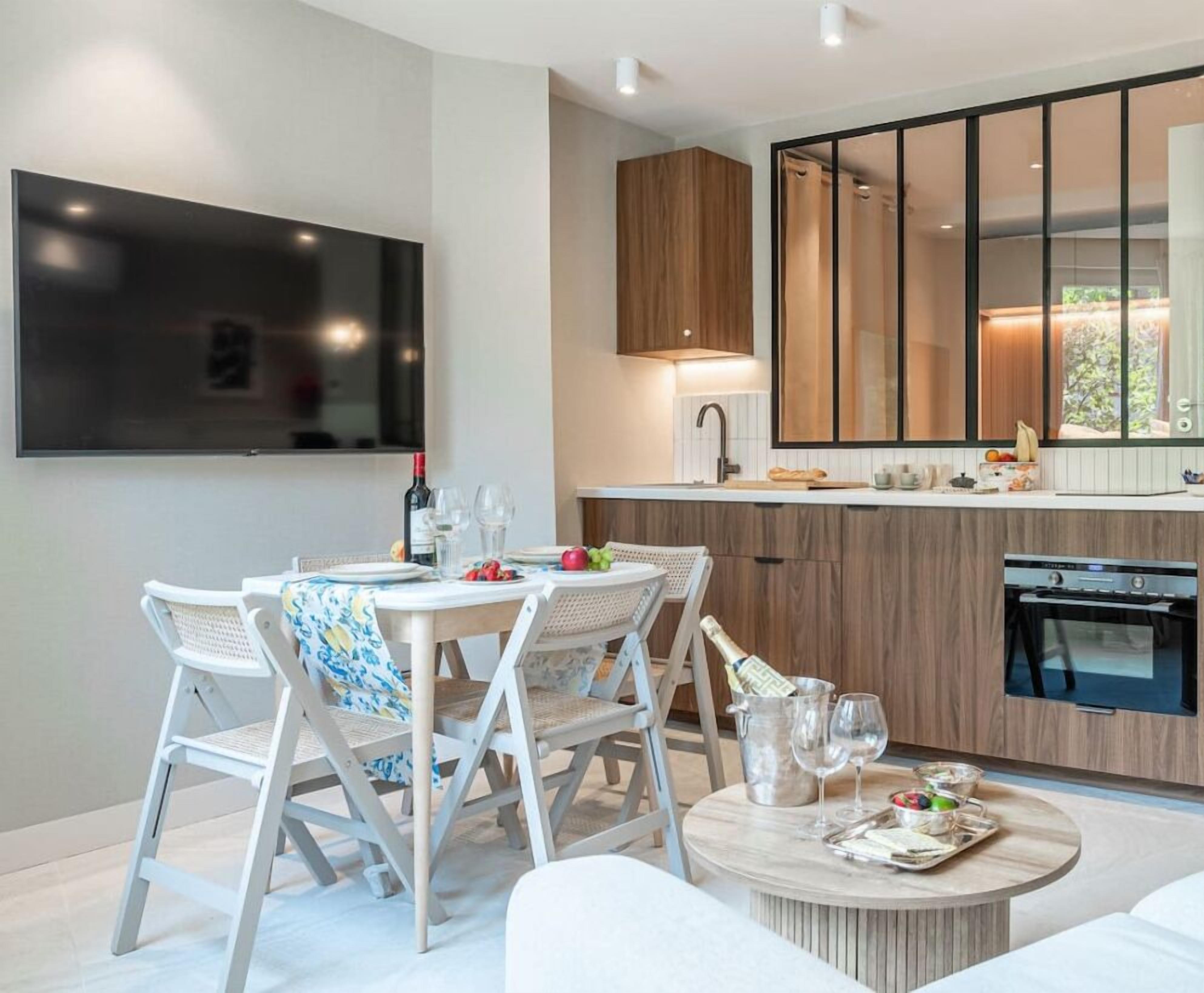 A modern kitchen with a TV, wooden cabinets, and a dining area with white chairs and a round table.
