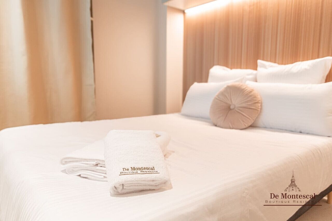 Una cama bien tendida con sábanas blancas y una toalla doblada, cabecera de madera y pared iluminada con luz cálida.