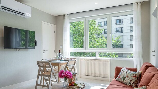 A modern living room with a dining area, large windows with a view of greenery, a flat-screen TV mounted on the wall, and a cozy seating area with a sofa. - De Montescal - Hot Tub A/C Wonder (Paris)