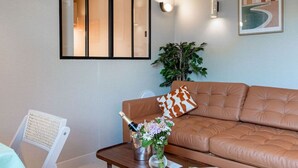 A living room with a brown leather sofa, a white chair, a wooden coffee table with a champagne bottle and flowers, and a framed picture on the wall.