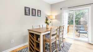 Un comedor con una mesa y sillas de madera, un florero con flores y una lámpara. Hay cuadros en la pared y se ve un balcón con una silla.