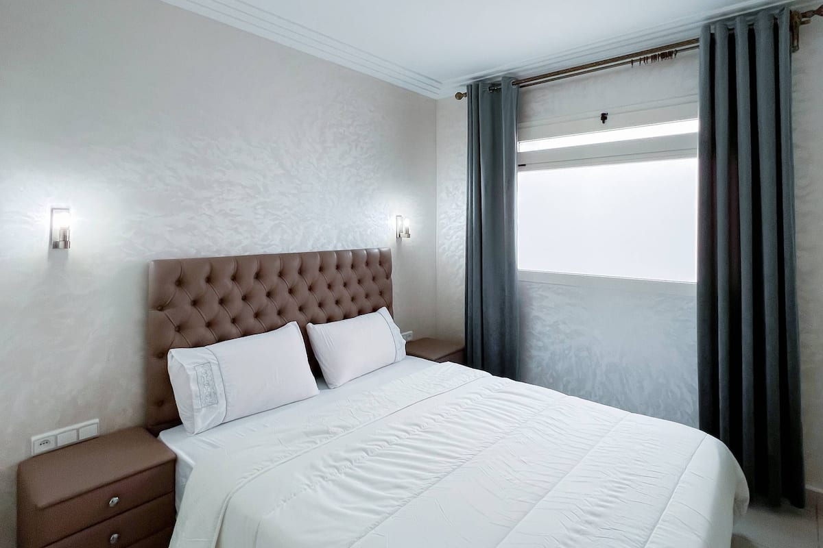 A modern hotel room with a large bed, a nightstand, and a window with curtains.