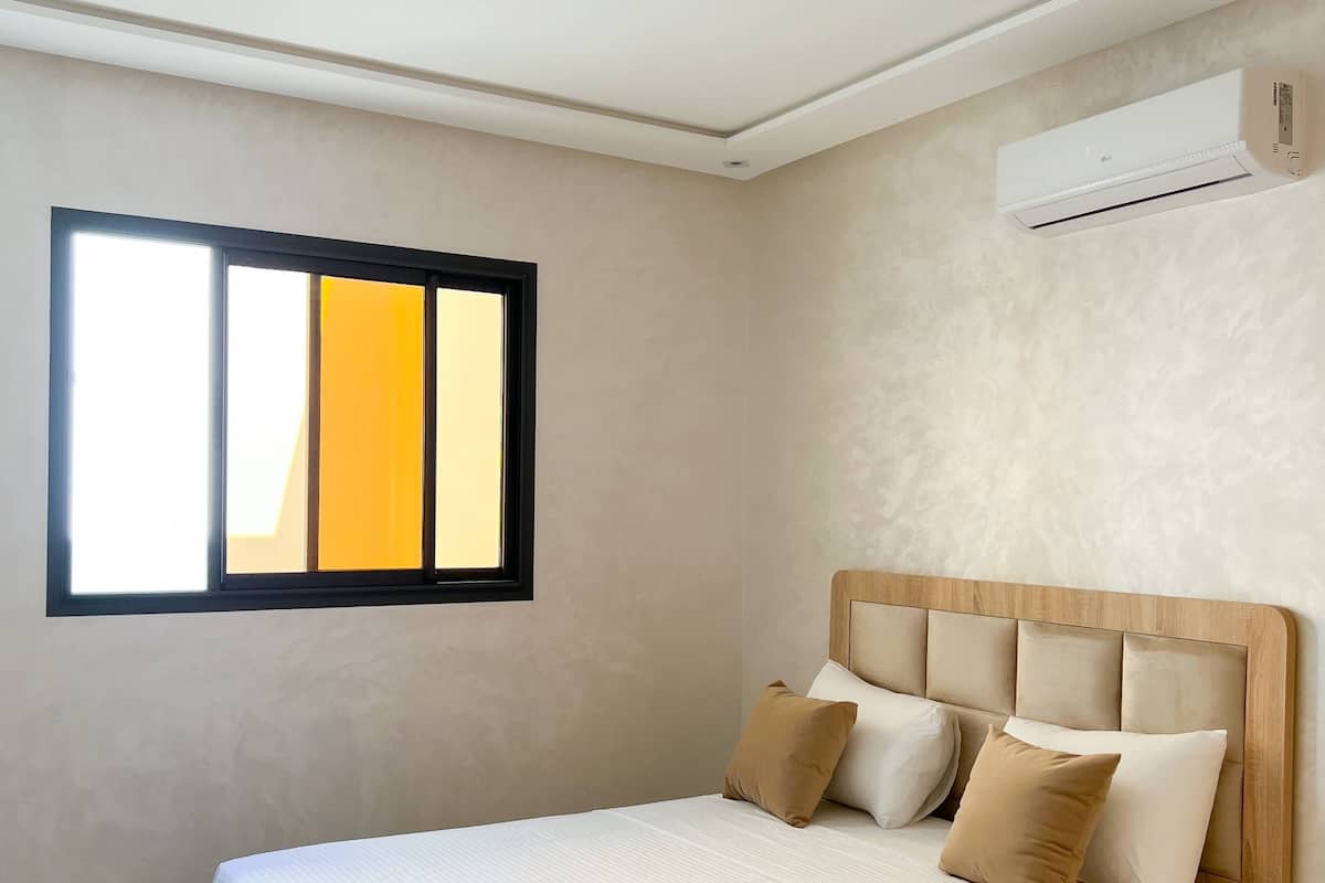 A bedroom with a bed, pillows, a window, and an air conditioning unit.