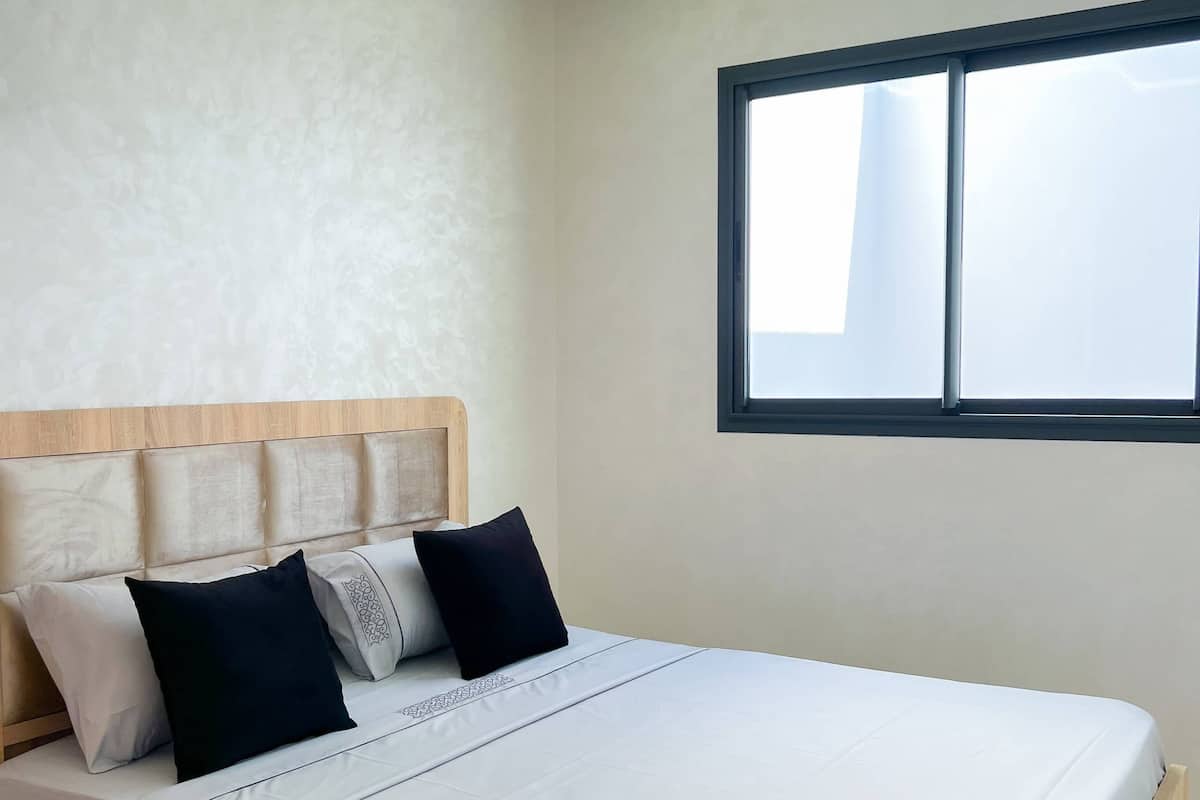 A modern bedroom with a wooden bed frame, a headboard with cushions, a large window, and recessed ceiling lighting.