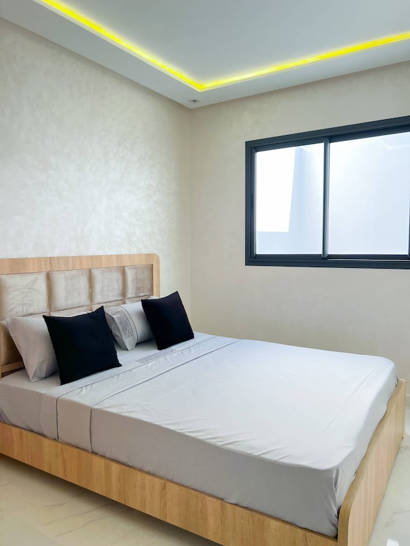 A modern bedroom with a wooden bed frame, a headboard with cushions, a large window, and recessed ceiling lighting. - Royal Rif Apartments (Ait Youssef Ou Ali)