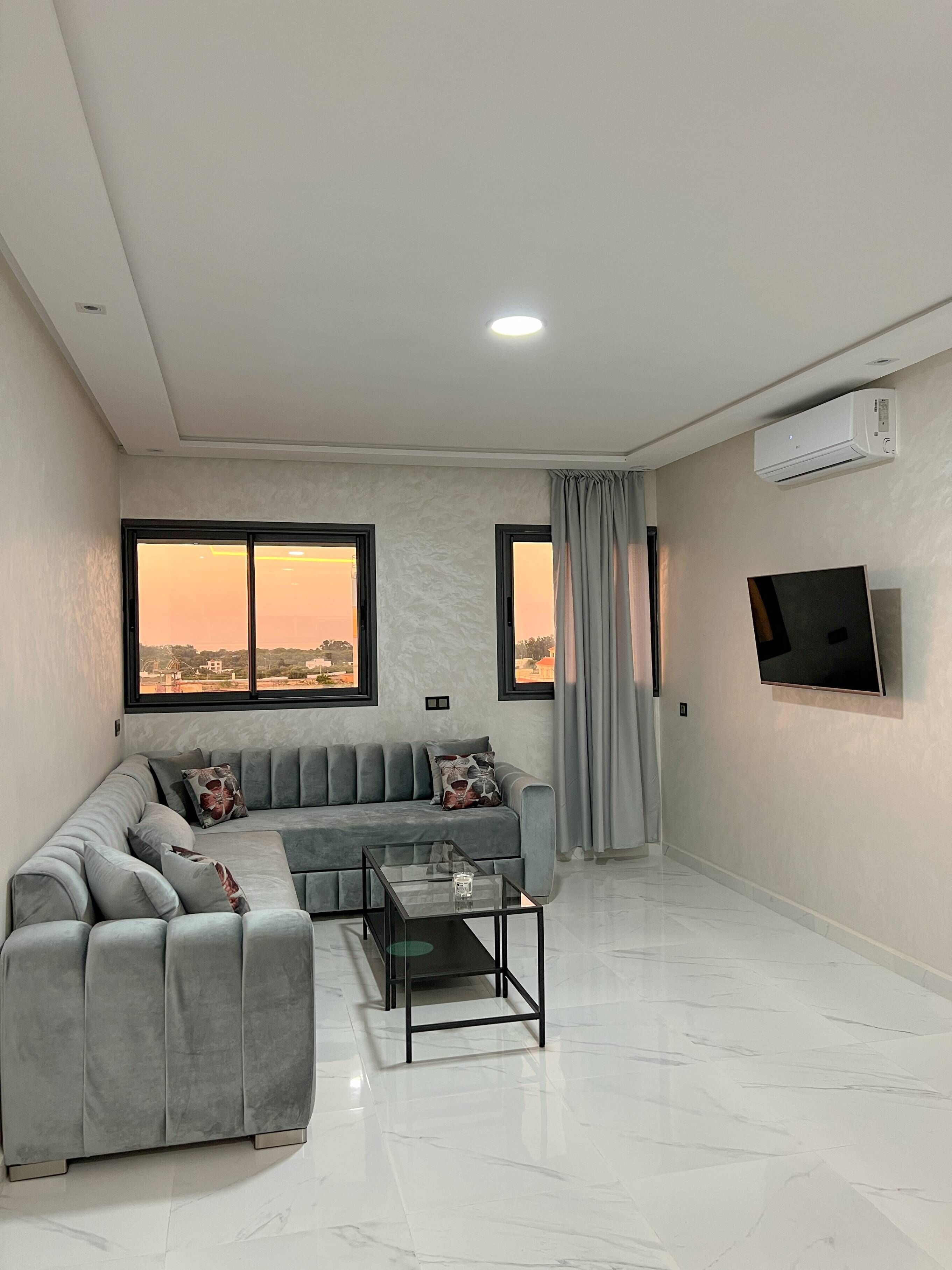 A modern living room with a grey sectional sofa, a glass coffee table, and a flat-screen TV mounted on the wall.