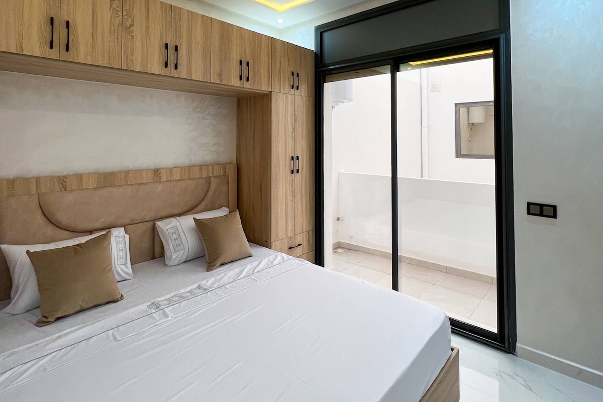 A modern bedroom with a large bed, wooden headboard, and a sliding glass door leading to a balcony.