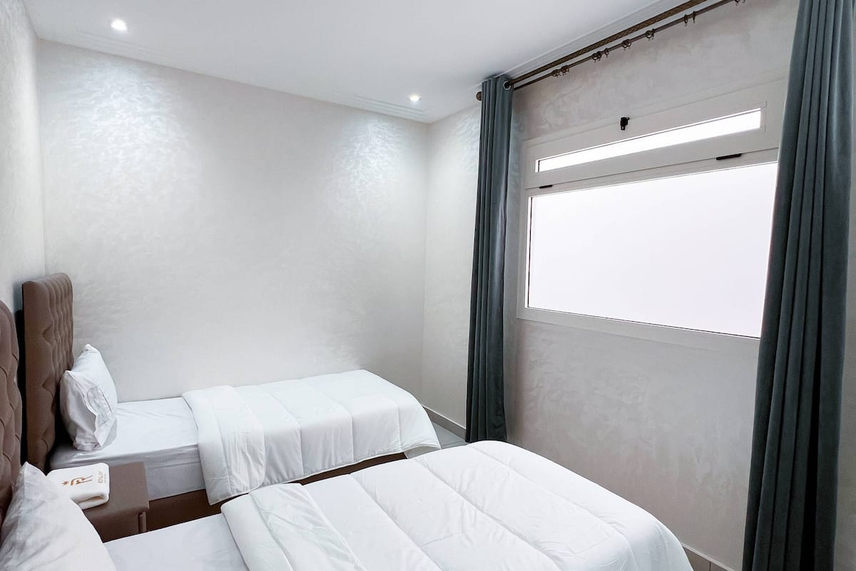 A hotel room with two beds, a window with curtains, and a small table with a lamp.