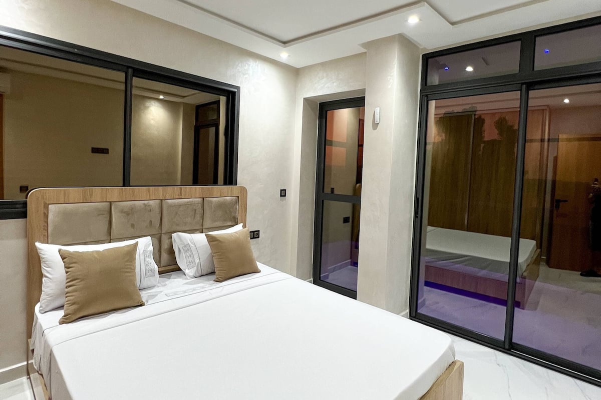 A modern bedroom with a large bed, a wooden headboard, and a sliding glass door leading to a balcony.