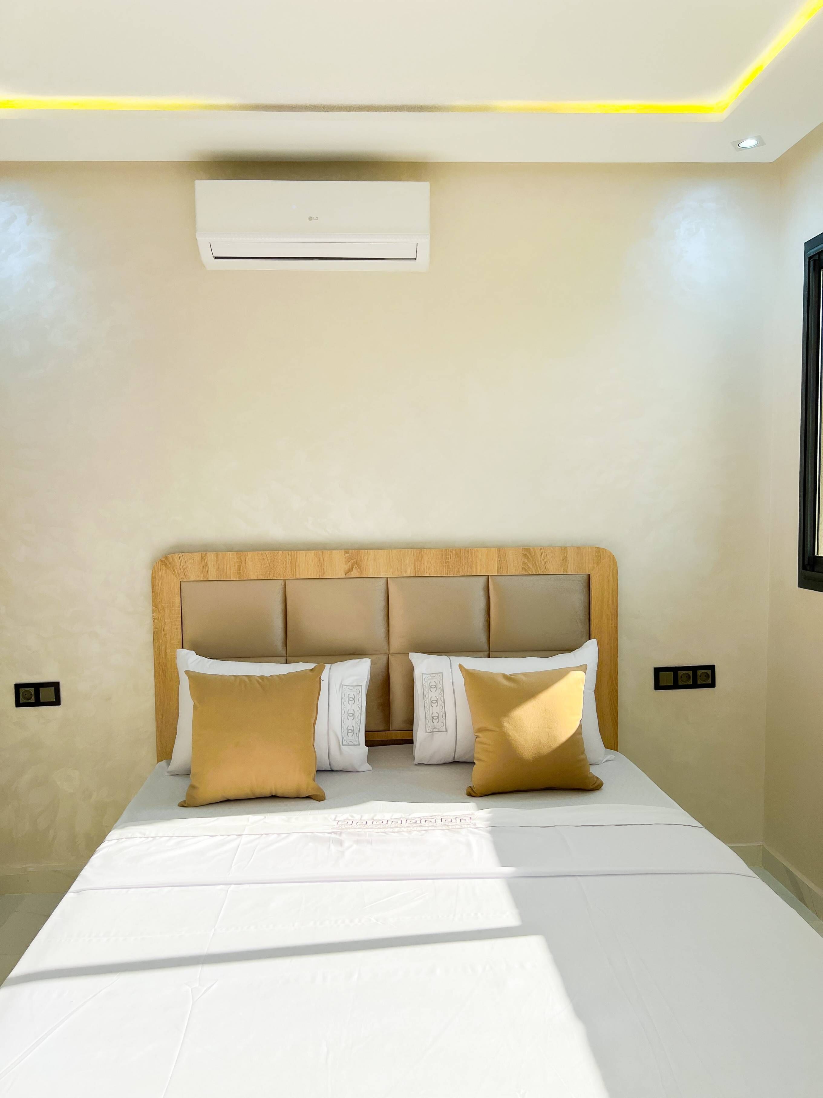 Un dormitorio con cabecera de madera, cama con ropa blanca, dos almohadas marrones y un equipo de aire acondicionado sobre la cama.