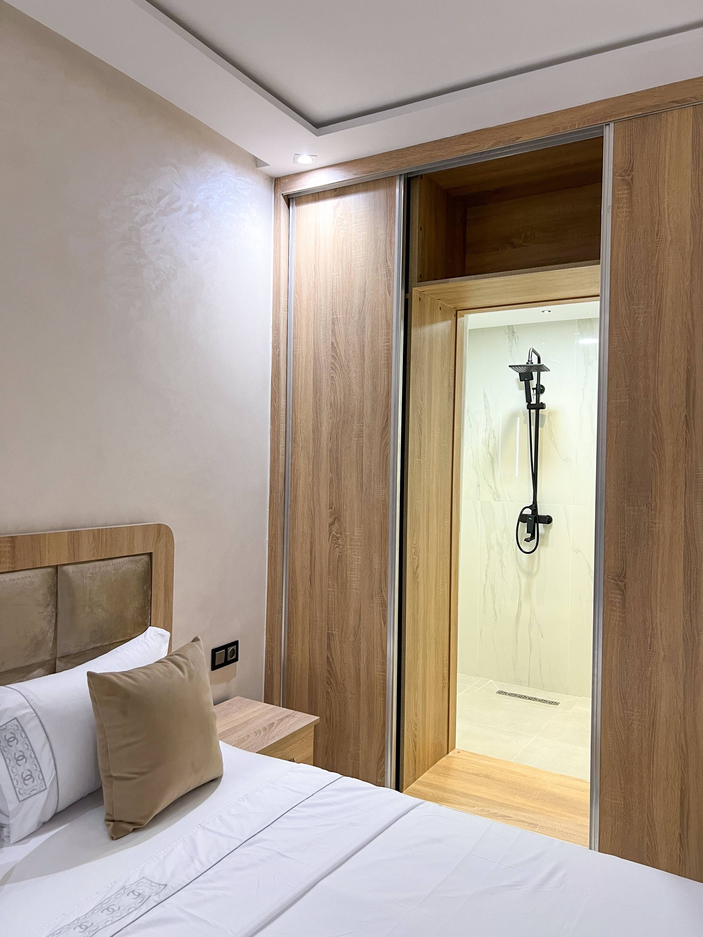 Habitación de hotel con cama, cabecera de madera, mesita de noche, almohada color beige y zona de ducha con mampara de cristal.