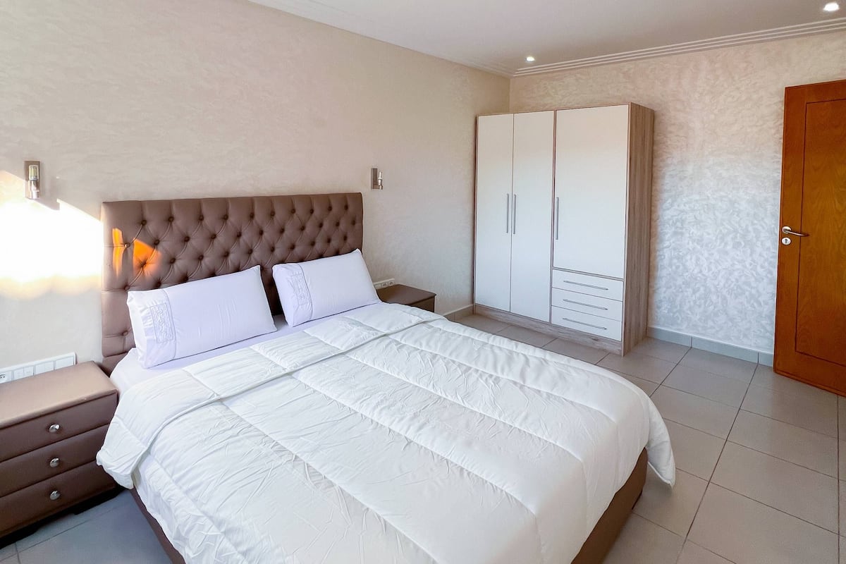 A neatly arranged bedroom with a large bed, a nightstand, a wardrobe, and a door.