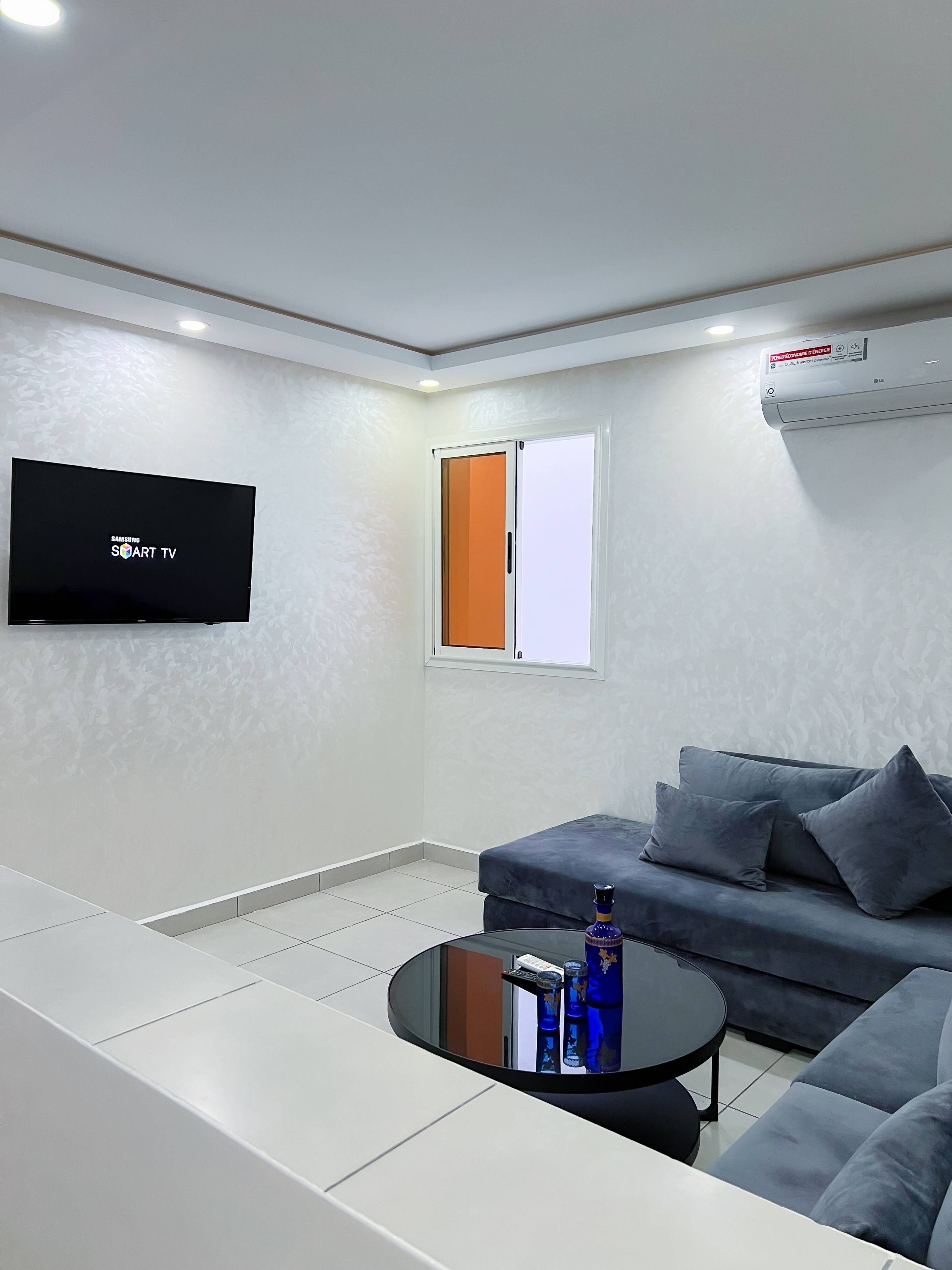 Una sala moderna con un televisor de pantalla plana montado en la pared, un sofá gris con cojines, una pequeña mesa redonda y una ventana con persiana naranja.