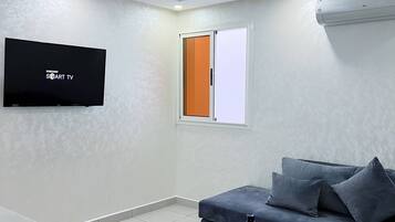 Una sala moderna con un televisor de pantalla plana montado en la pared, un sofá gris con cojines, una pequeña mesa redonda y una ventana con persiana naranja.