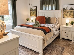 Une chambre à coucher avec un lit, des tables de chevet, une commode, une lampe, une plante et une fenêtre avec des rideaux.