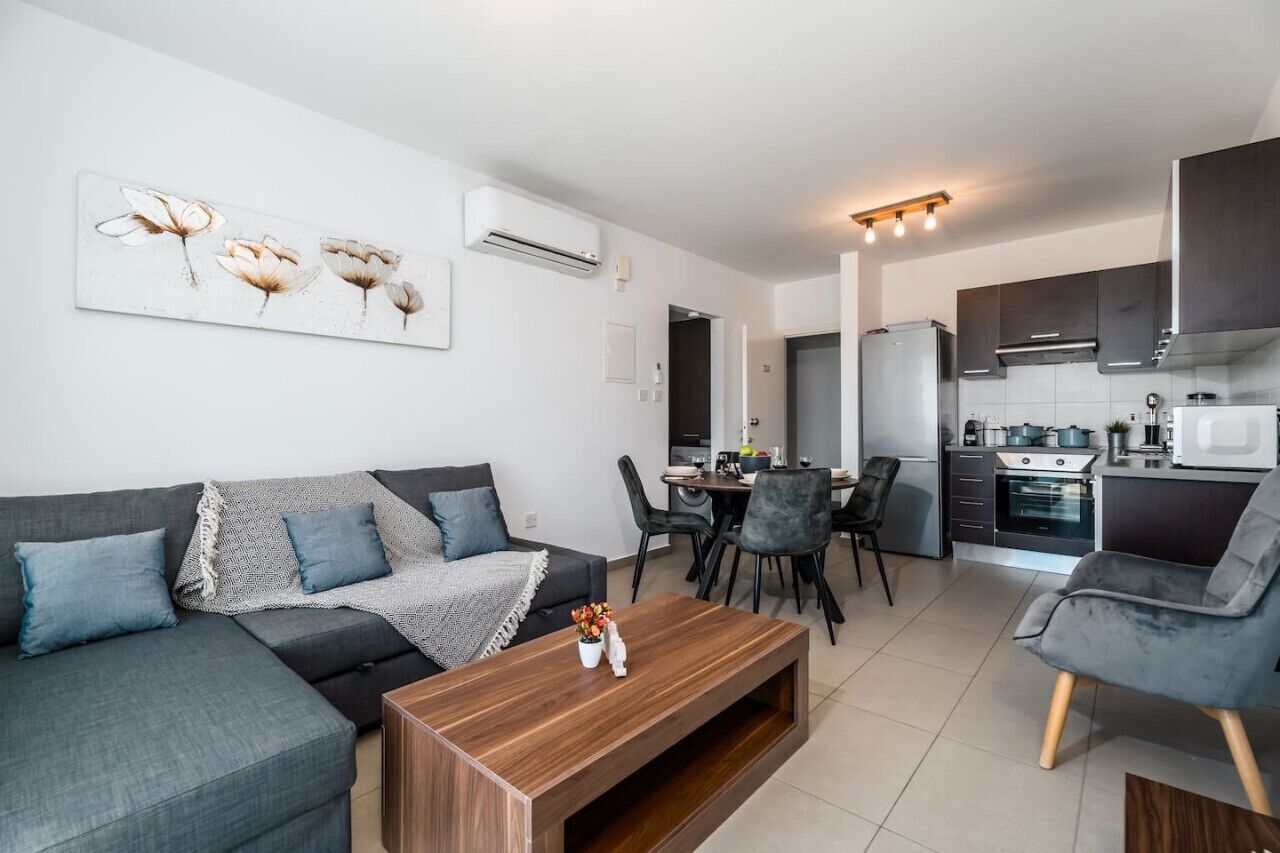 Una sala de estar moderna con un sofá gris, una mesa de centro de madera y un comedor con mesa y sillas. Se ve una cocina con gabinetes y electrodomésticos oscuros al fondo.