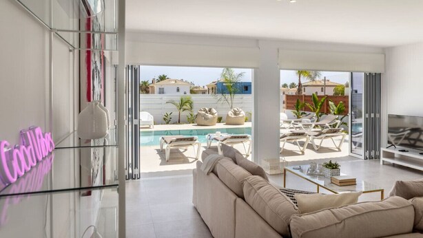 A modern living room with a large sofa, a glass coffee table, and a pool area visible through the glass doors. - SunnyVillas: 3BR New Modern Villa*Private Pool*BLG (Paralimni)