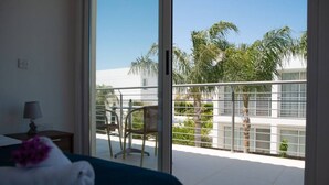 Une chambre d’hôtel avec un lit, une table de chevet, une lampe et une vue sur un balcon agrémenté de chaises et de palmiers.