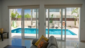 Un salon moderne avec un canapé, une table basse et une vue sur une piscine, visible à travers de grandes portes-fenêtres coulissantes.