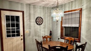 Comedor con mesa y sillas de madera, candelabro, dardo, ventana con persianas y puerta con ventanal.