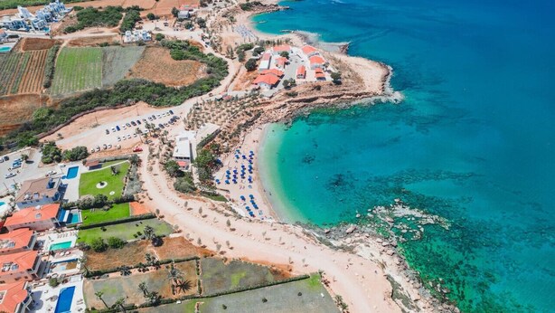 Vue aérienne d’une zone côtière avec une plage, des bâtiments et une eau d’un bleu limpide.