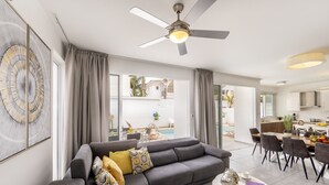 Un salon moderne avec un canapé gris, un coin repas avec une table blanche et une vue sur une piscine et un espace extérieur.