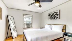 Une chambre à coucher avec un lit, un ventilateur de plafond, une fenêtre et une œuvre d’art encadrée accrochée au mur.