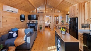 Ein Interieur einer Holzhütte mit Holzbalkendecke, einer Couch, einer Küchenzeile inklusive Kühlschrank und einem Esstisch.