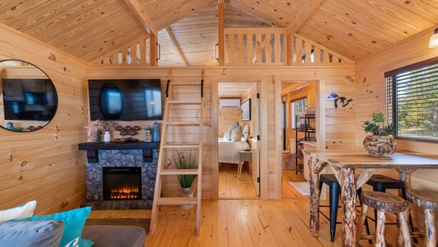 Ein gemütliches Holzhäuschen mit Kamin, einem Essbereich mit rustikalen Hocker und einem Wohnzimmer mit Sofa und Kissen.
