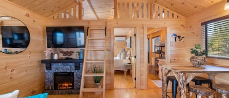 L’intérieur d’un chalet en bois chaleureux et accueillant, avec une cheminée, un coin repas doté de tabourets rustiques et un salon avec un canapé et des coussins.