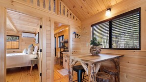 Ein Holzhütten-Innenraum mit einem Bett, einem Esstisch mit Hocker und einem großen Fenster mit Jalousien.