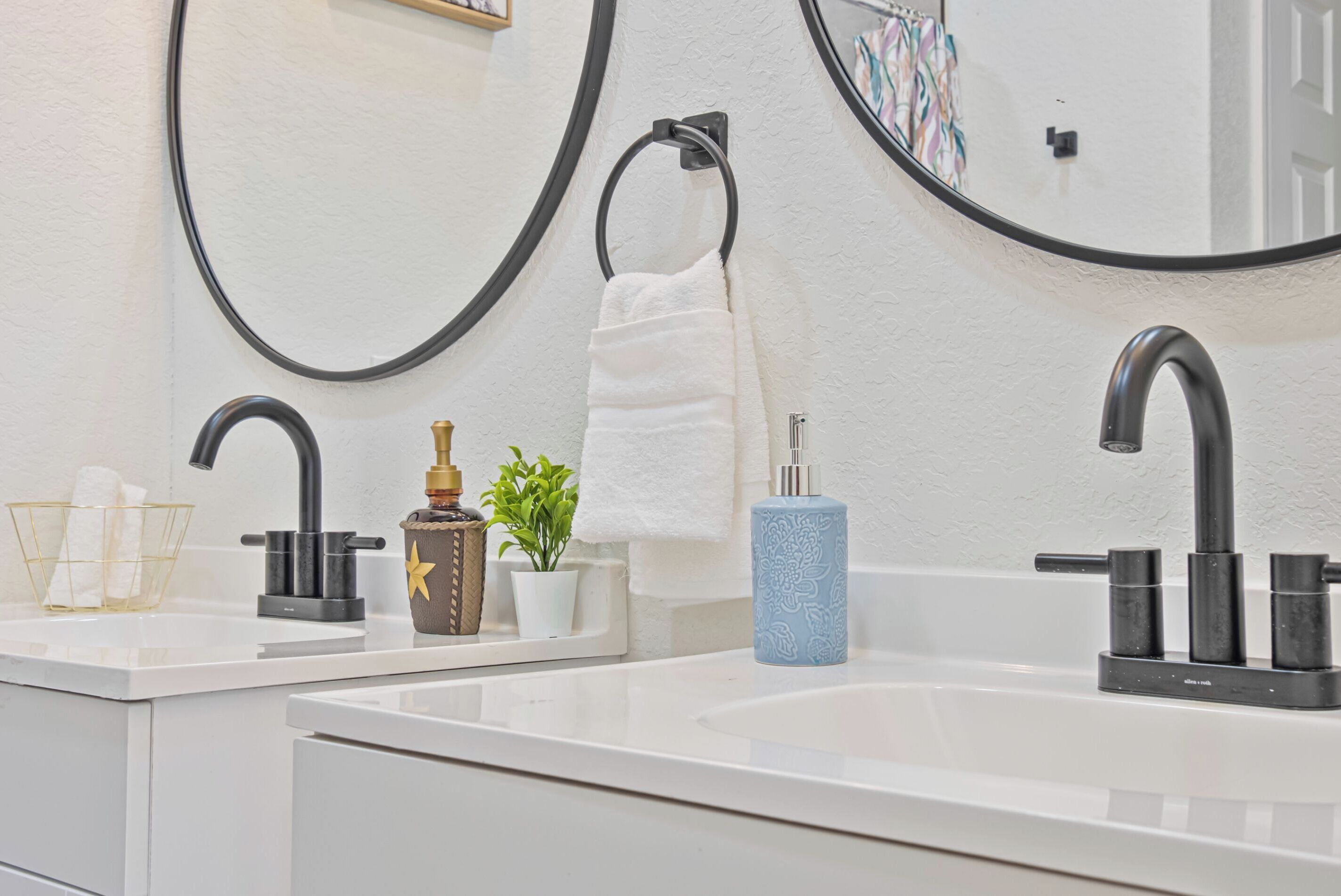Uma casa de banho moderna com duas pias, torneiras pretas, uma toalha branca, um dispensador de sabonete azul e uma pequena planta em vaso.
