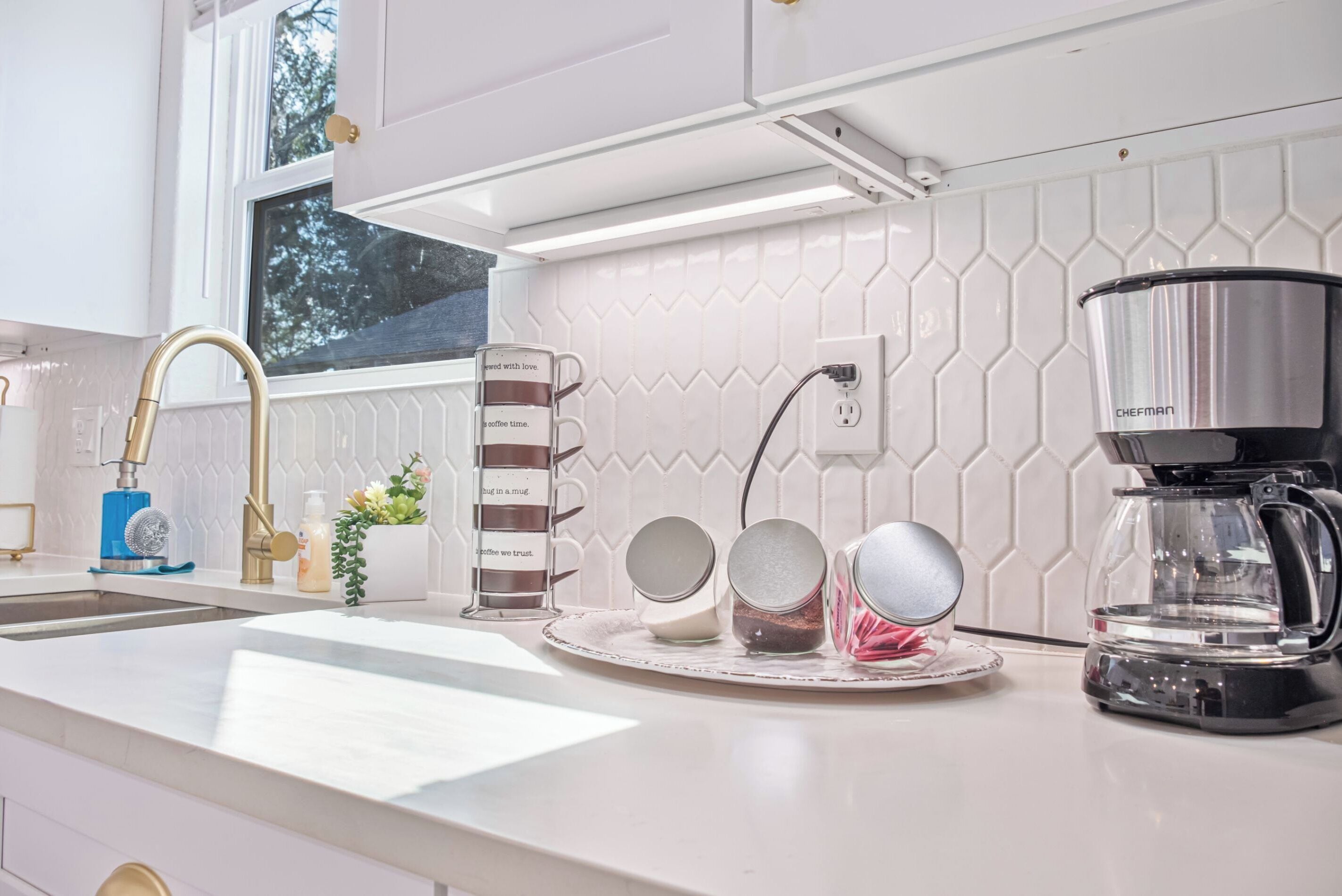 A modern kitchen with white cabinetry, a stainless steel coffee maker, and a window providing natural light.