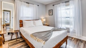 A neatly arranged bedroom with a wooden bed, white bedding, a bedside table with a lamp, and sheer curtains. - Short Walk to Wesley Hospital | 3 Beds W/ W&D! (Wichita)